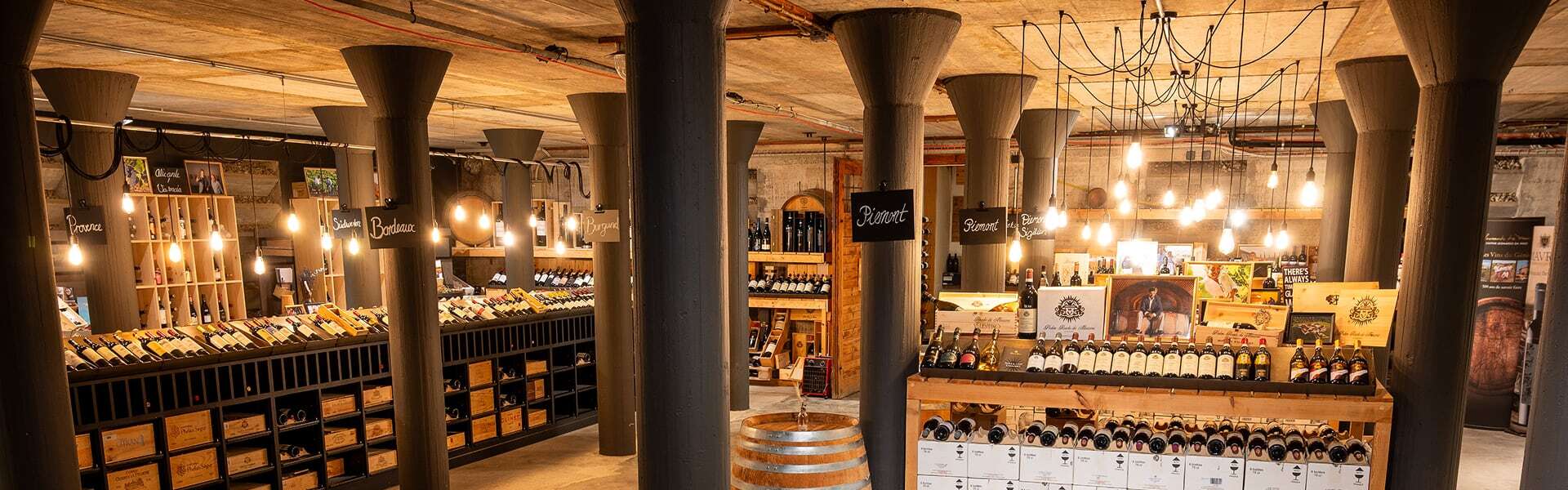Großer Verkaufsraum mit mehreren Säulen, langen Regalen voller Weinflaschen und warmen Hängelampen über den Auslagen