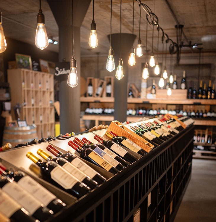 Schräg gelagerte Weinflaschen stehen in langen Regalen unter hängenden Glühbirnen in einem Verkaufsraum.
