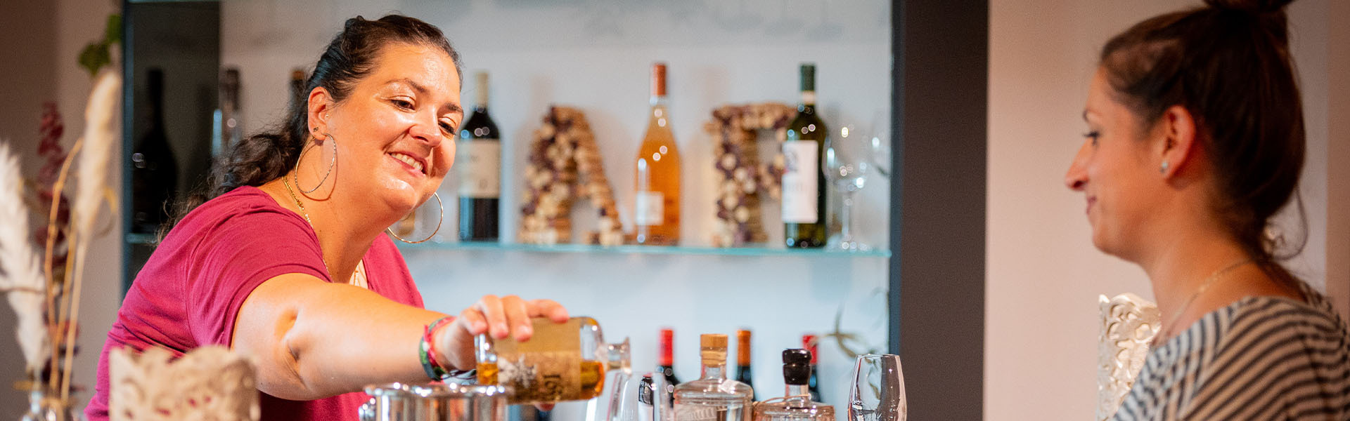 Eine lächelnde Mitarbeiterin gießt einen Whiskey in ein Glas an der Bar, eine Kundin steht ihr gegenüber.