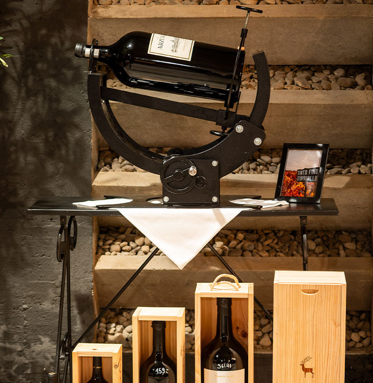 Eine große Weinflasche liegt in einer mechanischen Vintage-Weinwaage auf einem Tisch, darunter stehen mehrere Flaschen in Holzkisten.